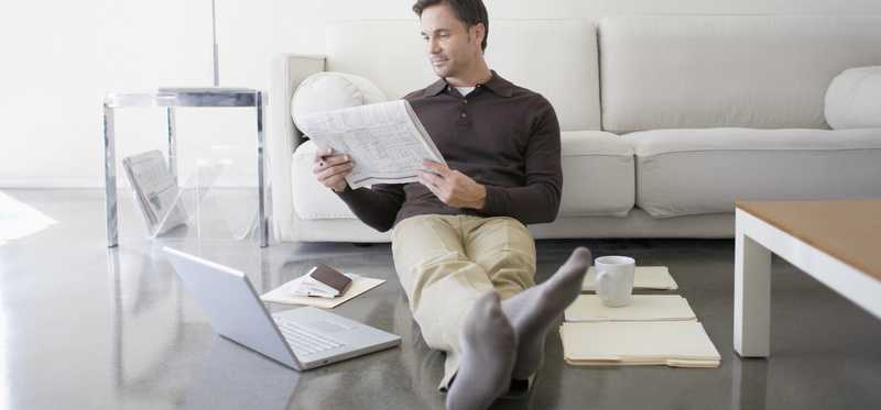 Man reading newspaper while sitting on floor with laptop and folders nearby