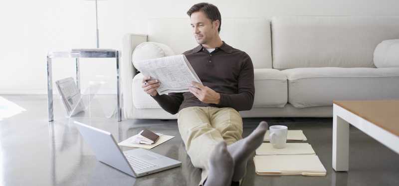Man reading newspaper while sitting on floor with laptop and folders nearby