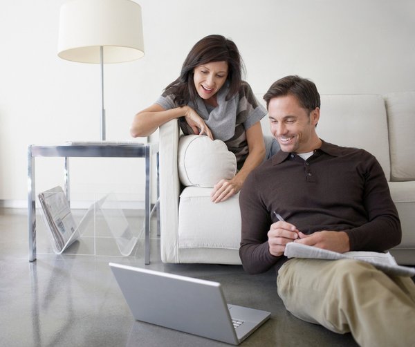 One person sitting on a couch and another sitting on the floor next to them. Both are smiling while looking at a laptop also sitting on the floor.