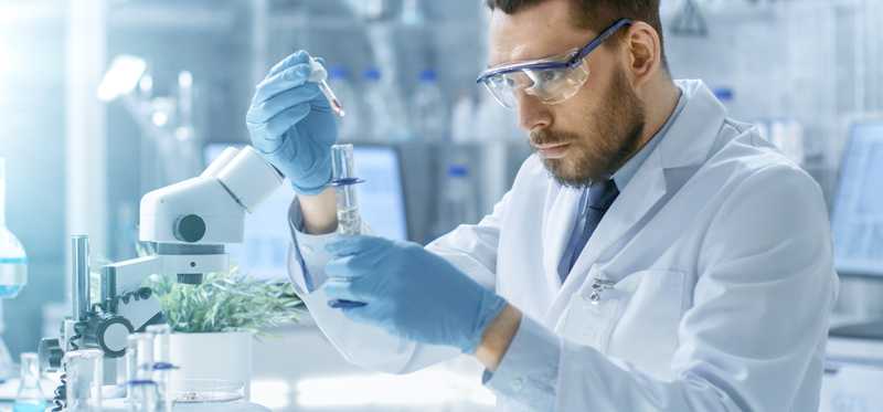 Man in lab attire holding a dropper over a test tube.