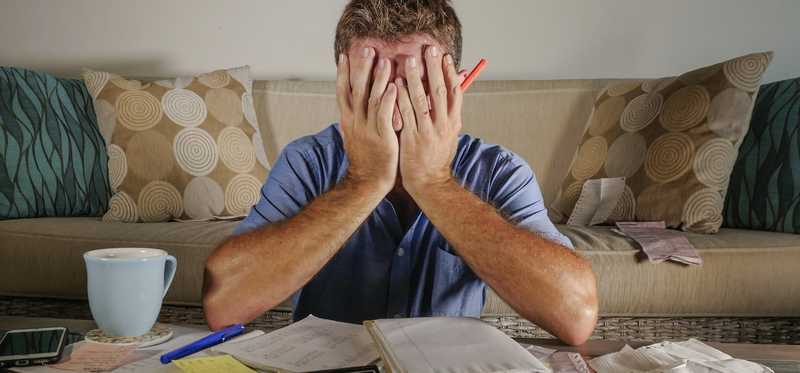 Person covering face behind table covered in papers, a calculator, a notebook, a pen, a mug, and a mobile phone.