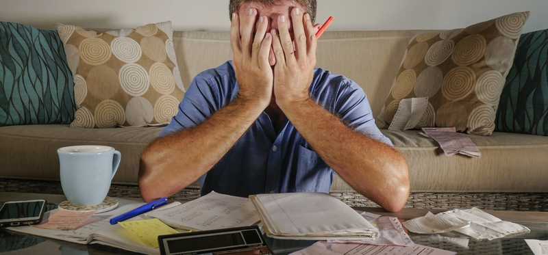 Man covering face behind table covered in papers, a calculator, a notebook, a pen, a mug, and a mobile phone