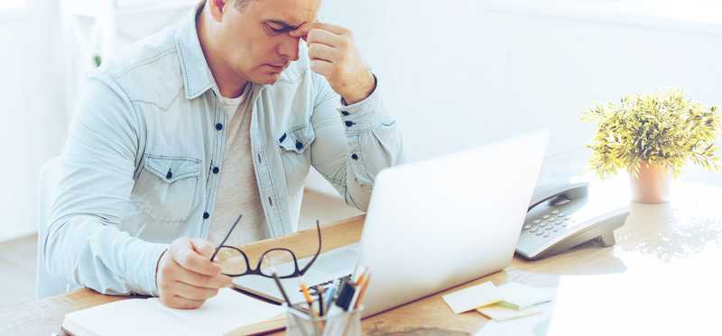 Mature man sitting at laptop with paperwork pinching the bridge of his nose in frustration.