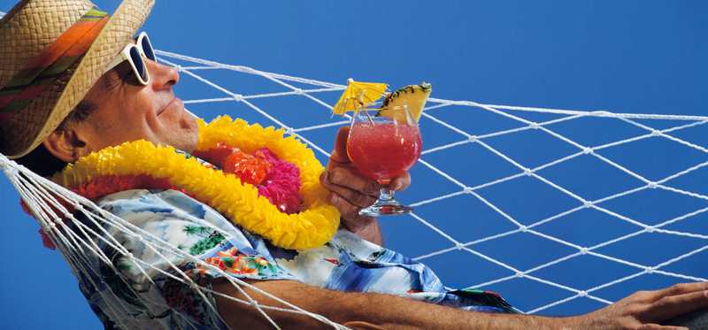 Person in a sun hat and lei lying in a hammock with drink.
