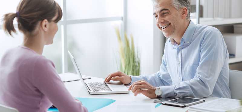Smiling person at laptop sitting across from another person at desk.