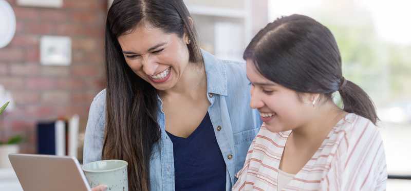 Smiling adult stands by smiling teenager who reviews information on laptop.