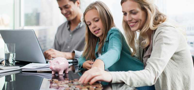 An adult and child count coins from a piggy bank while another adult uses a laptop.