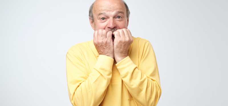An older man in yellow shirt bites his fingernails.