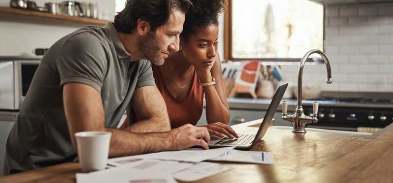A couple sitting at their kitchen counter looking at a laptop.