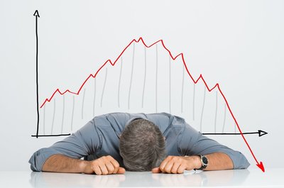 Man with head on table in front of falling stock chart.