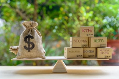 A balance with a money bag on one side and stacked boxes of ETFs, stocks, bonds, and other assets on the other side.