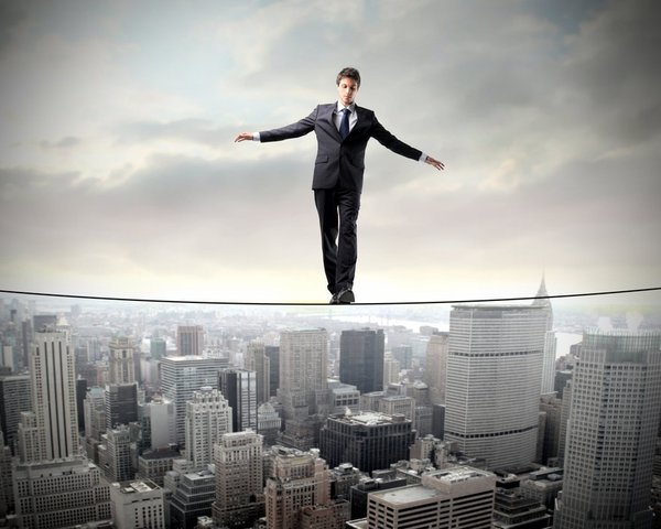 Business man in suit balances on a tightrope above a cloudy city