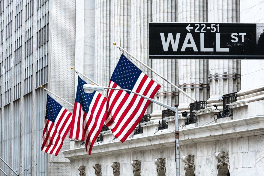 The exterior of an old building with three American flags and the Wall St. street sign in the foreground.