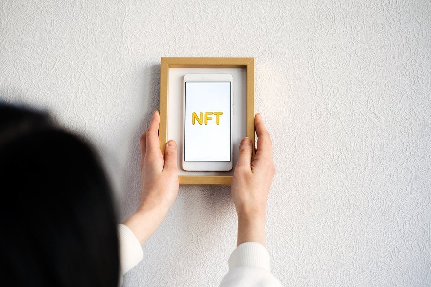 A woman placing a picture frame over a phone displaying the word NFT.