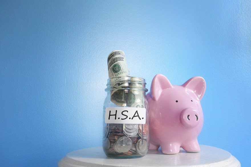 Glass jar full of coins and bills with H.S.A. on label next to a piggy bank.