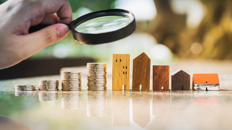 A person holding a magnifying glass looking at a row of rising coins and buildings.