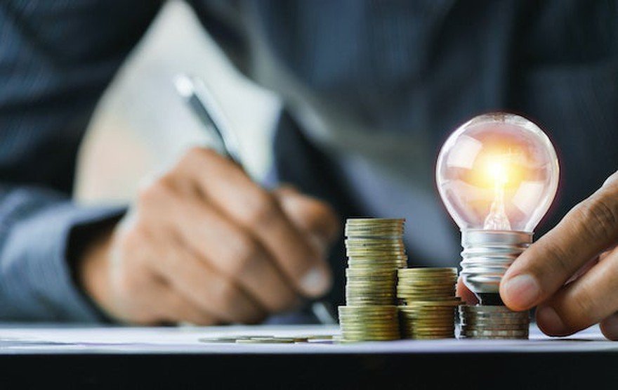 man writing on paper holding a lit up lightbulb above a stack of coins