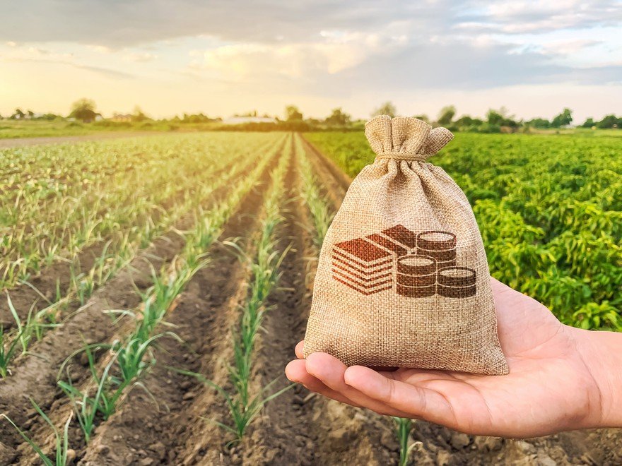 A hand holding a money bag with a farm in the background.