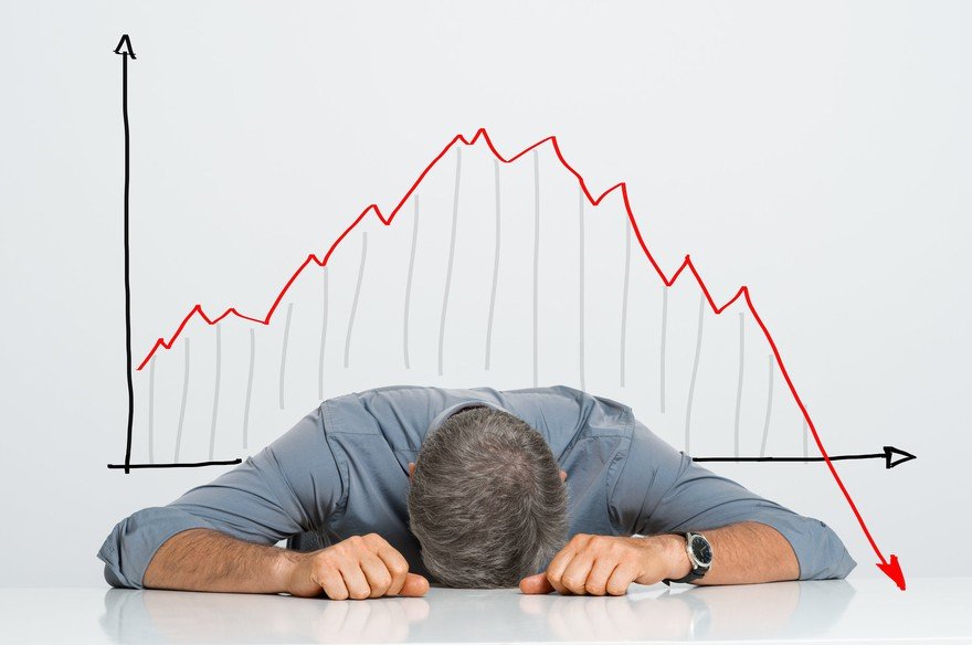 An investor lying their head on a table in front of a falling stock chart.