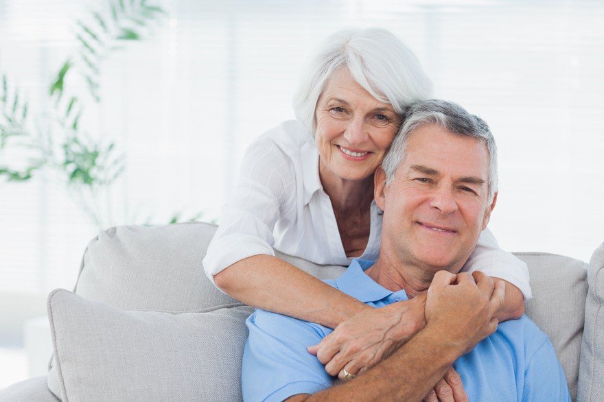 Grinning mature woman leans over a couch, holding an older smiling male. 