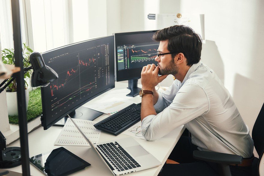 Back view of bearded stock market broker in eyeglasses analyzing data and graphs on multiple computer screens while sitting in modern office. Stock exchange. Trade concept. Investment concept