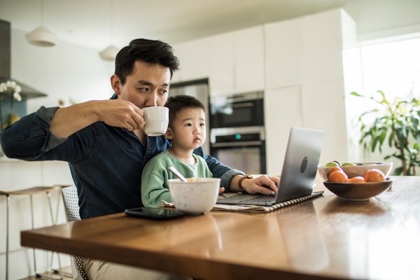 Person drinking from coffee mug while using laptop and holding child on lap.