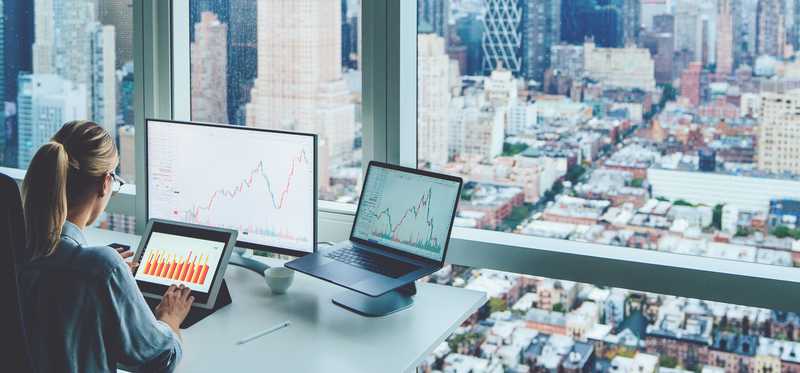 A person at a desk with many monitors and a big office window overlooking a big city.