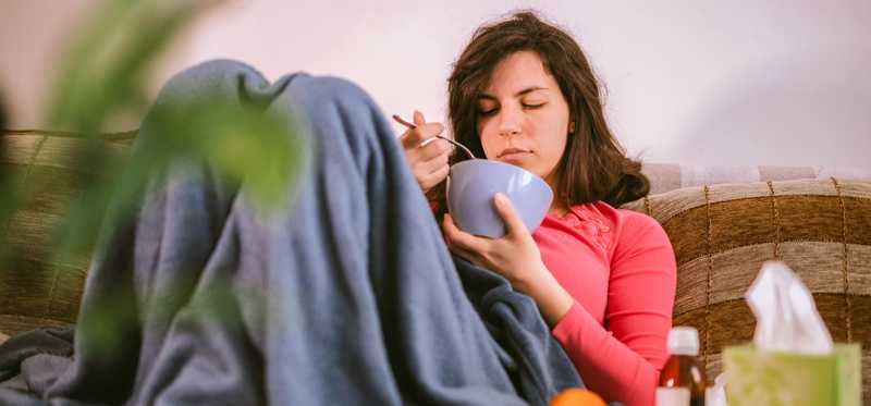 Person eating a bowl of soup while wrapped in a blanket.