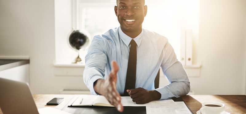 Smiling business professional sitting at a desk and extending a handshake to unseen individual.