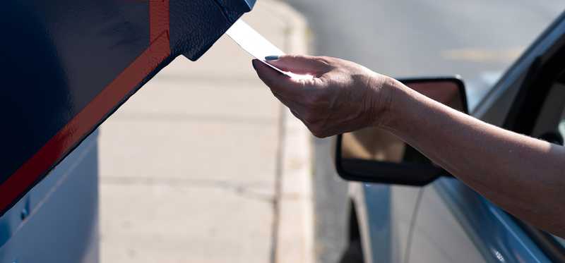 A hand putting mail in a mail slot outside a post office.