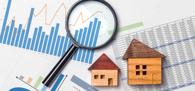 Two small wooden houses and a magnifying glass lying atop investing charts and spreadsheets.
