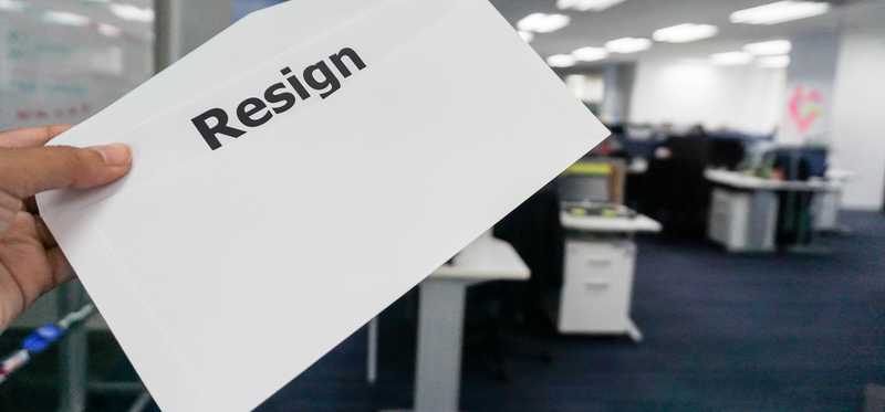Hand holding piece of paper with the word Resign on it in an office