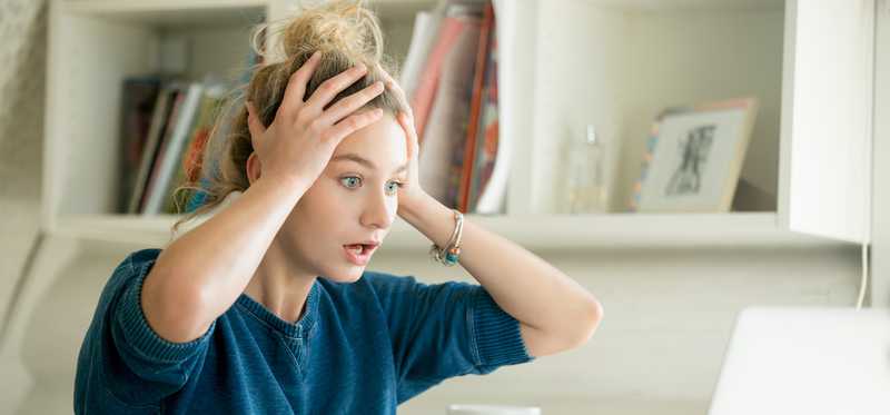 Woman in front of laptop with hands on her head looking shocked