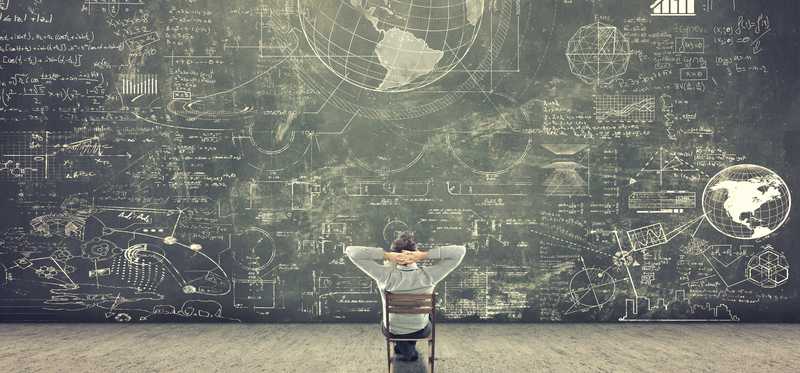 Man sitting in chair with hands behind head admiring a chalkboard wall full of drawings and info.