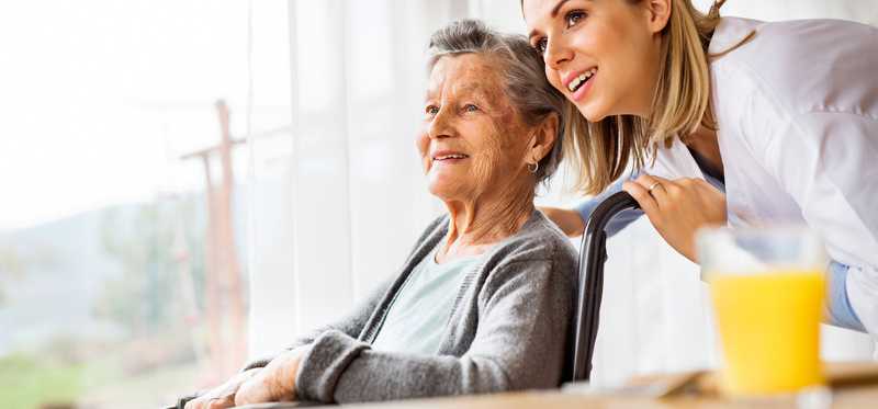 Young woman leans down to talk to elderly woman in wheelchair.