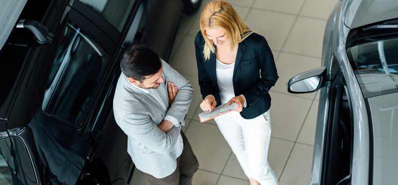 A woman and man stand between two cars on a showroom floor.