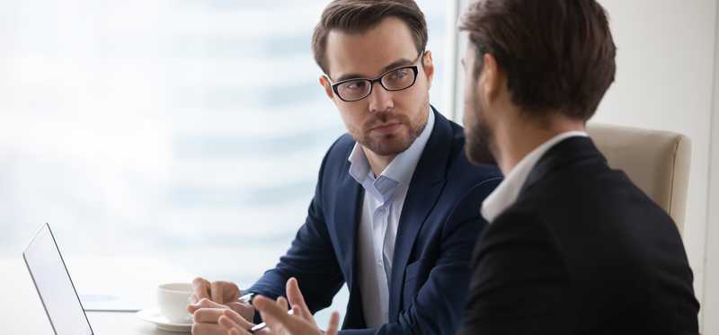 Two businessmen sit at desk with laptop and chat.