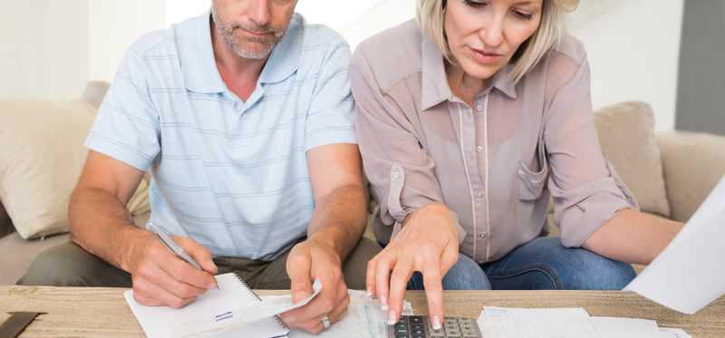 Adult couple sits on couch and looks over paperwork with a calculator.