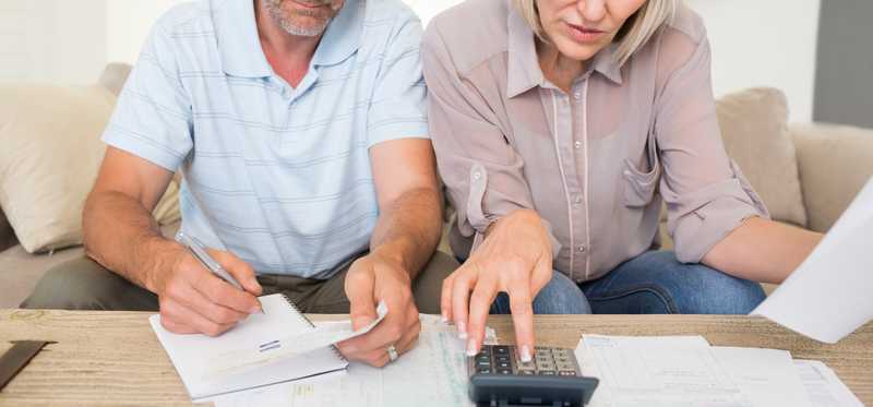 Adult couple sits on couch and looks over paperwork with a calculator.