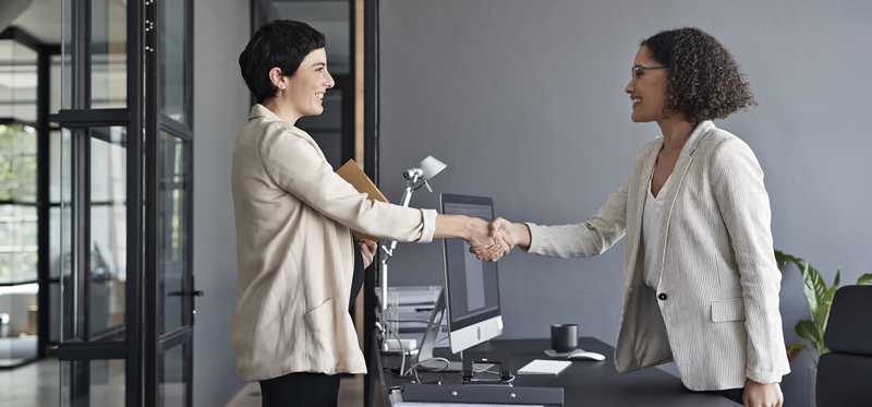Two businesspeople standing and shaking hands over a desk.