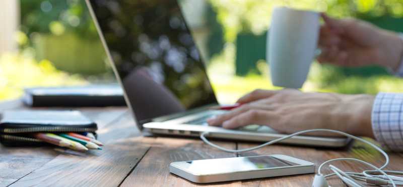 Hands typing on a laptop with a cell phone and headphones nearby.