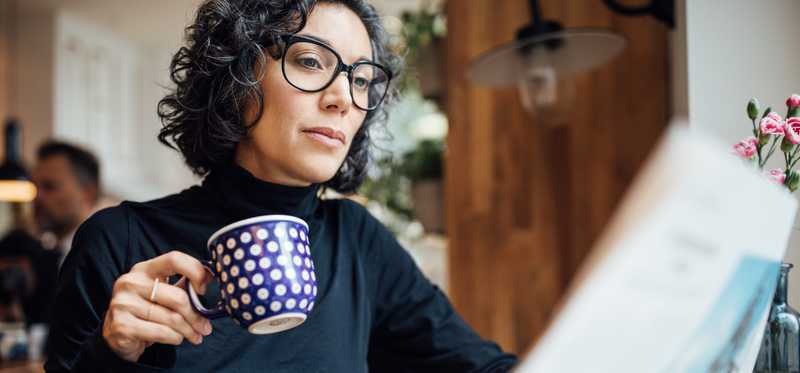 Person in glasses drinking from coffee mug and reading the newspaper.