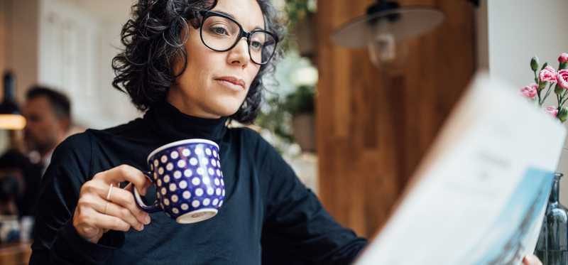 Person in glasses drinking from coffee mug and reading the newspaper.