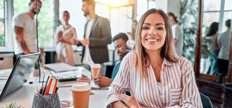 Person smiling and sitting at table with cup of coffee and other people around.