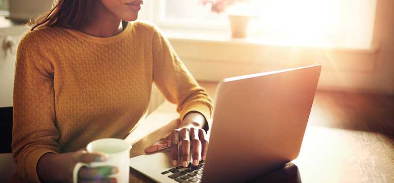 A woman on a laptop with a cup of coffee, in front of a sunny window.
