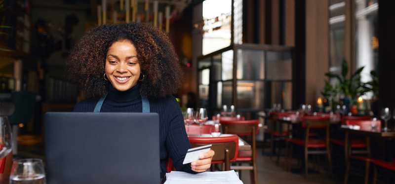 A person working in a café and holding a credit card while processing a customer's payment at a laptop.