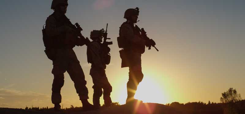 Soldiers in uniform with weapons backlit by sunset.