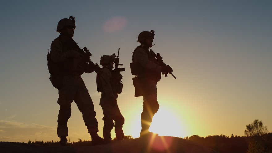 Soldiers in uniform with weapons backlit by sunset.