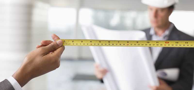 Person holding a tape measure in a blank space and construction worker in background.