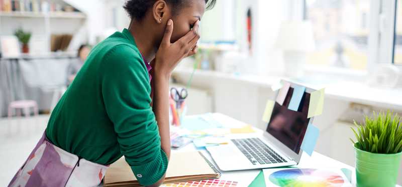 Stressed young woman sitting at desk in front of laptop with her face in her hands